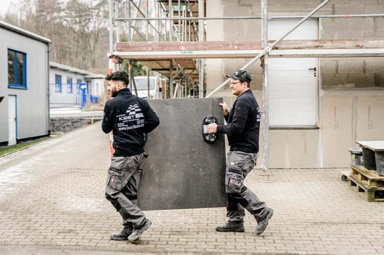 Gemeinsam tragen zwei Handwerker vom Fliesenleger Meisterbetrieb Fliesen Kast aus Heimbach eine Großformatfliese auf einer Baustelle in Düren