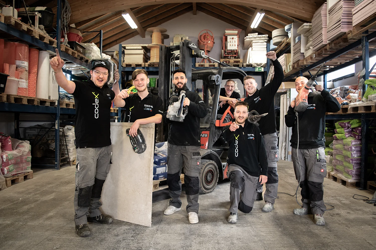 Das Team von Fliesen Kast steht in einer Lagerhalle und jubelt mit Werkzeug und Material.